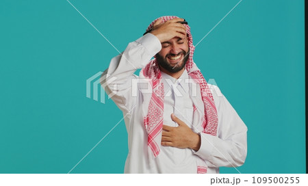 Cheerful person laughing at a joke in studio, having fun while he wears muslim traditional clothing and kufiyah. Young arab man enjoying laugh with people, expressing happiness and joy. 109500255