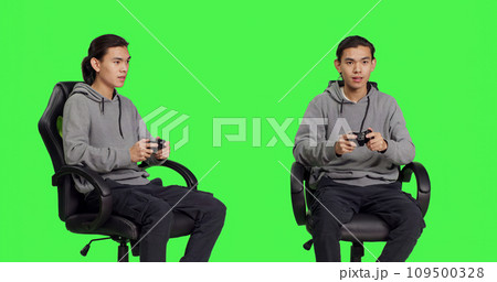 Asian guy playing videogames over greenscreen backdrop, using controller and having fun with friends on online gaming competition. Young man gamer enjoying rpg gameplay, blank background. Asian guy playing videogames over greenscreen backdrop, using controller and having fun with friends on online gaming competition. Young man gamer enjoying rpg gameplay, blank background. 109500328