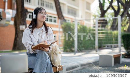 A positive female is listening to music and keeping her diary while resting on a bench in a park. 109506397
