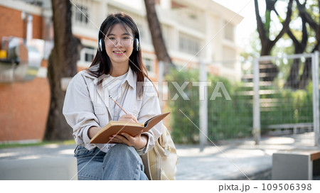 A positive female is listening to music and keeping her diary while resting on a bench in a park. 109506398