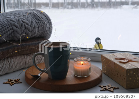 Winter windowsill still life. Blue ceramic cup of hot coffee on window sill. Christmas decorations on the background. Cozy home picture. Warm woolen knitted sweaters, Burn Candle, Cookies. Stock photo 109509507