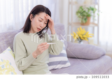 体調不良　体温計を見ながら頭痛で痛い頭をおさえるミドル女性 109511033