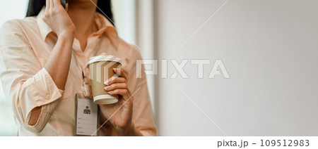 Young woman employee holding a paper cup of hot coffee and talking on mobile phone Young woman employee holding a paper cup of hot coffee and talking on mobile phone 109512983