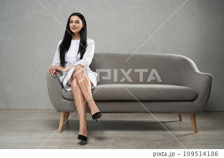 Attractive caucasian female medical staff looking at camera, while sitting on comfortable grey sofa. Front view of smiling woman wearing white robe posing with crossed legs, copy space. Work concept. Attractive caucasian female medical staff looking at camera, while sitting on comfortable grey sofa. Front view of smiling woman wearing white robe posing with crossed legs, copy space. Work concept. 109514186