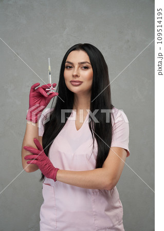 Confident female beautician wearing pink uniform standing with syringe indoors. Portrait of beautiful caucasian woman wearing gloves holding syringe and smiling in clinic. Concept of beauty care. Confident female beautician wearing pink uniform standing with syringe indoors. Portrait of beautiful caucasian woman wearing gloves holding syringe and smiling in clinic. Concept of beauty care. 109514195