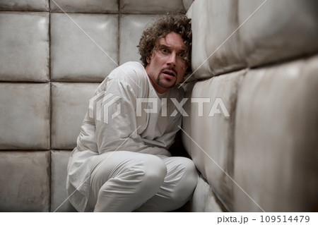Mad scared man patient wearing straitjacket in corner of padded room 109514479