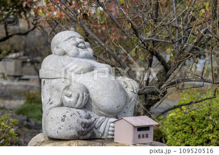Nagahama, Shiga Prefecture, Japan. Nov 21, 2023. A sitting smiling Buddha stone figure at a Japanese a Shinto shrine. Nagahama, Shiga Prefecture, Japan. Nov 21, 2023. A sitting smiling Buddha stone figure at a Japanese a Shinto shrine. 109520516
