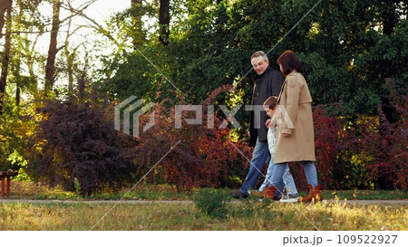 Family with son spends weekend together walking across fall botanical garden. Family of mom and dad leads little son by hands along path with bushes and bench. Family in warm clothes enjoy warm sunset Family with son spends weekend together walking across fall botanical garden. Family of mom and dad leads little son by hands along path with bushes and bench. Family in warm clothes enjoy warm sunset 109522927