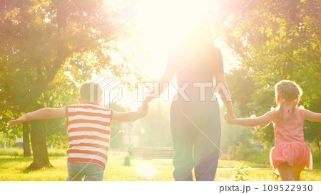 Silhouette of mother with hands interlocked with children leading across park lawn. Mother and children run across park with hands interlocked in bond of shared joy. Mother leads children through park 109522930