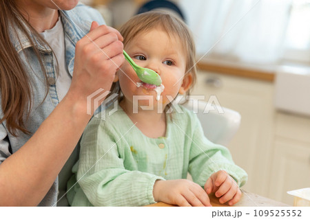 Happy family at home. Mother feeding her baby girl from spoon in kitchen. Little toddler child with messy funny face eats healthy food at home. Young woman mom giving food to kid daughter Happy family at home. Mother feeding her baby girl from spoon in kitchen. Little toddler child with messy funny face eats healthy food at home. Young woman mom giving food to kid daughter 109525552