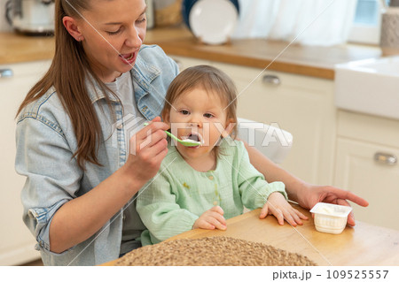 Happy family at home. Mother feeding her baby girl from spoon in kitchen. Little toddler child with messy funny face eats healthy food at home. Young woman mom giving food to kid daughter Happy family at home. Mother feeding her baby girl from spoon in kitchen. Little toddler child with messy funny face eats healthy food at home. Young woman mom giving food to kid daughter 109525557