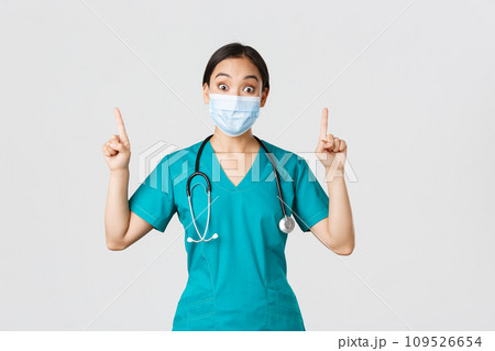 Covid-19, coronavirus disease, healthcare workers concept. Intrigued asian female doctor, physician or intern in medical mask and scrubs, looking curious, pointing fingers up, white background 109526654