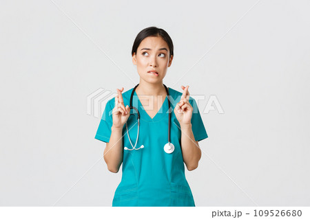 Covid-19, healthcare workers, pandemic concept. Hopeful nervous asian female intern in scrubs waiting for news, doctor cross fingers and biting lip as looking upper left corner, praying 109526680