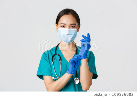Covid-19, coronavirus disease, healthcare workers concept. Professional smiling asian female physician, doctor in medical mask and scrubs, put on rubber gloves ro examination, white background Covid-19, coronavirus disease, healthcare workers concept. Professional smiling asian female physician, doctor in medical mask and scrubs, put on rubber gloves ro examination, white background 109526682