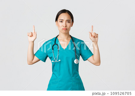 Covid-19, healthcare workers, pandemic concept. Gloomy disappointed asian female doctor, physician in scrubs pointing fingers up and grimacing displeased, standing white background Covid-19, healthcare workers, pandemic concept. Gloomy disappointed asian female doctor, physician in scrubs pointing fingers up and grimacing displeased, standing white background 109526705