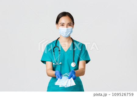 Covid-19, coronavirus disease, healthcare workers concept. Friendly smiling asian physician, female doctor in scrubs and rubber gloves handing over medical masks to patients, white background 109526759
