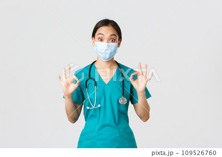 Covid-19, coronavirus disease, healthcare workers concept. Excited and impressed asian female doctor, intern in medical mask and scrubs showing okay gesture in approval, white background 109526760