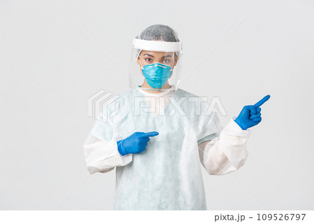 Covid-19, coronavirus disease, healthcare workers concept. Professional female doctor, tech lab employee in personal protective equipment pointing fingers right, showing way, white background Covid-19, coronavirus disease, healthcare workers concept. Professional female doctor, tech lab employee in personal protective equipment pointing fingers right, showing way, white background 109526797