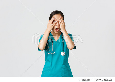 Covid-19, healthcare workers and preventing virus concept. Scared and shocked asian female nurse, doctor in scrubs witness something embarrassing or scary, cover eyes but peek through fingers 109526838