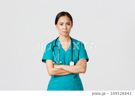 Covid-19, healthcare workers, pandemic concept. Skeptical and reluctant asian female doctor, tired nurse in scrubs cross arms and frowning, smirk displeased, standing white background 109526842