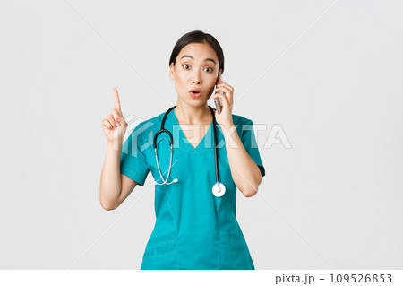 Covid-19, healthcare workers and preventing virus concept. Portrait of beautiful asian female doctor, nurse in scrubs talking on phone and raise finger as have great idea, remember, suggest plan 109526853