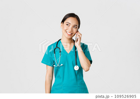 Covid-19, healthcare workers and preventing virus concept. Pretty smiling asian female doctor, physician in scrubs having conversation, talking on phone and looking upper left corner dreamy 109526905