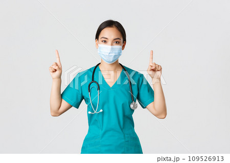 Covid-19, coronavirus disease, healthcare workers concept. Young professional asian female doctor, nurse in medical mask and scrubs, pointing fingers up, showing way, advertising 109526913