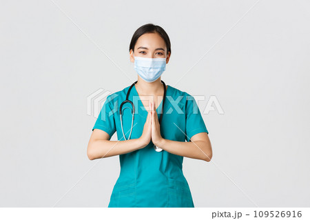 Covid-19, coronavirus disease, healthcare workers concept. Hopeful smiling asian female physician, doctor in medical mask and scrubs hold hands in praying gesture, thanking, white background 109526916