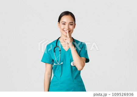 Covid-19, healthcare workers, pandemic concept. Cheerful, smiling female asian nurse in scrubs having secret, making surprise, showing shush gesture, shhh keep quiet, standing white background 109526936