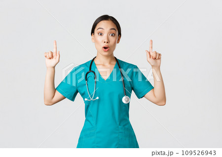 Covid-19, healthcare workers, pandemic concept. Surprised and thrilled asian female nurse, woman doctor in scrubs asking question interesting promo, pointing fingers up, showing clinic advertisement 109526943