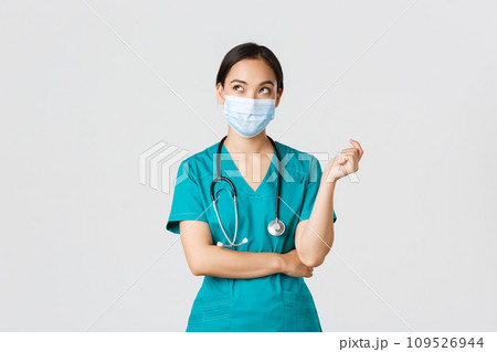 Covid-19, coronavirus disease, healthcare workers concept. Curious and thoughtful asian female doctor in medical mask and scrubs, looking upper left corner, thinking, white background 109526944
