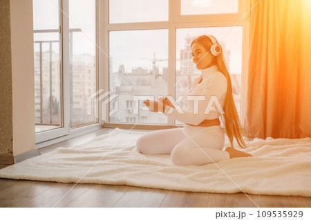 Side view portrait of relaxed woman listening to music with headphones lying on carpet at home. She is dressed in a white tracksuit. Side view portrait of relaxed woman listening to music with headphones lying on carpet at home. She is dressed in a white tracksuit. 109535929