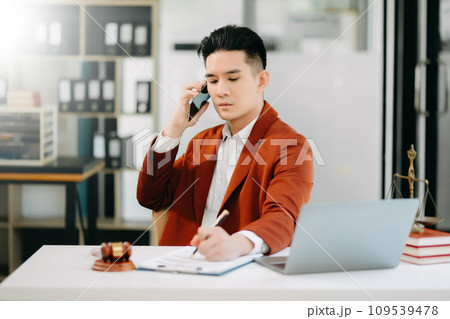 Asian male lawyer chatting online with client on laptop, smartphone  discussing details of lawsuit. in office 109539478