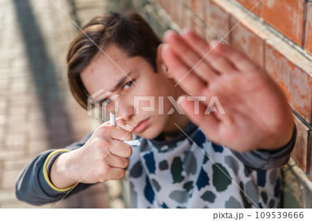 A caucasian teenage boy holds a broken cigarette in his hand and shows a stop sign, protesting against smoking. The concept of adolescent smoking addiction 109539666