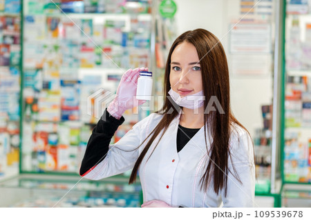 A pharmacist in a medical mask and gloves lowered to her chin,holding a jar of medicine in her hand.In the background, the pharmacy Windows are blurred.Concept of protection against virus and medicine A pharmacist in a medical mask and gloves lowered to her chin,holding a jar of medicine in her hand.In the background, the pharmacy Windows are blurred.Concept of protection against virus and medicine 109539678