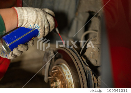 A man is repairing a car in a garage. A man treats rusted bolts with WD-40 solution. Replacing a brake disc on a car in the garage. Concept of a man fixing his car in a garage. 109541051