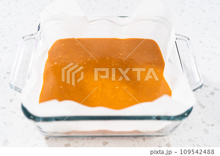 Pouring fudge mixture into the square glass baking pan lined with parchment paper to prepare pumpkin spice fudge. Pouring fudge mixture into the square glass baking pan lined with parchment paper to prepare pumpkin spice fudge. 109542488