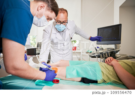 Two doctors making ultrasound diagnostics and injection in knee at hospital. Male specialists looking, examining patient, boy lying on couch. Concept of medicine and healthcare. 109556470