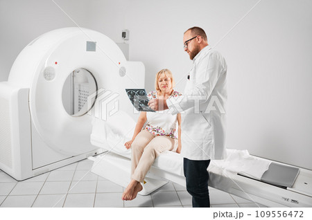 Medical computed tomography or MRI scanner. Doctor holding and explaining results of MRI. Woman patient after MRI, sitting on couchette. Concept of medicine, healthcare and modern diagnostics. 109556472