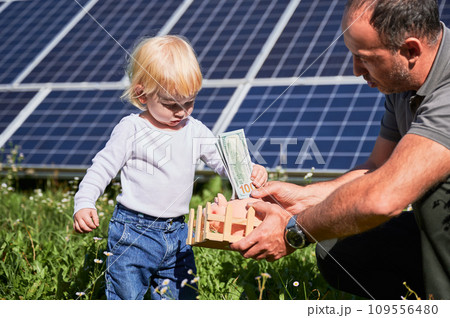Son and his father putting cash into piggy bank together on background of solar panels. Dad explaining his kid importance of saving money for future. Man and his child investing money to piggy bank 109556480