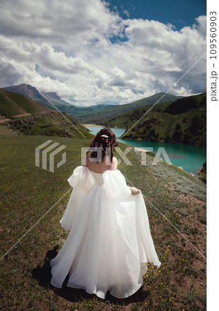 beautiful bride in a white dress stands or walks in the autumn mountains among green grass against the backdrop of a lake in the distance. Scenic wedding photography 109560903