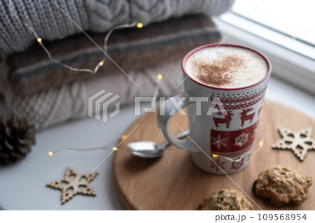 Winter windowsill still life. Red ceramic cup of hot coffee on window sill. Christmas decorations on the background. Cozy home picture. Warm woolen knitted sweaters, Burn Candle, Cookies. Stock photo Winter windowsill still life. Red ceramic cup of hot coffee on window sill. Christmas decorations on the background. Cozy home picture. Warm woolen knitted sweaters, Burn Candle, Cookies. Stock photo 109568954