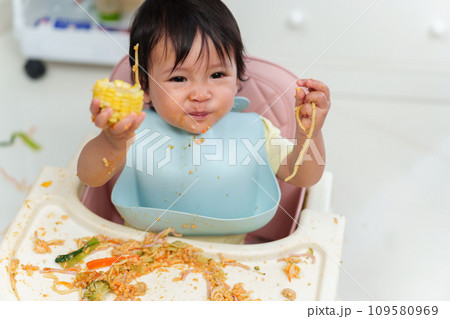 happy infant baby eating food and vegetable by self feeding BLW or baby led weaning on chair happy infant baby eating food and vegetable by self feeding BLW or baby led weaning on chair 109580969