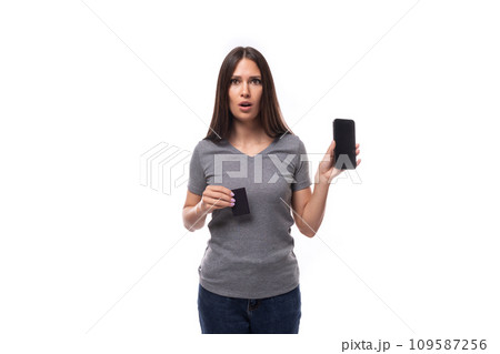 young european brunette woman dressed in a gray basic t-shirt holds a smartphone and a plastic card 109587256