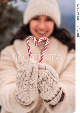 Woman candy sea. Smiling woman in knitted hat, mittens and beige coat holding lollipops candy canes in her hands in shape of heart against the backdrop of the sea. Woman candy sea. Smiling woman in knitted hat, mittens and beige coat holding lollipops candy canes in her hands in shape of heart against the backdrop of the sea. 109587429