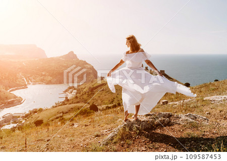 Happy woman in a white dress and hat stands on a rocky cliff above the sea, with the beautiful silhouette of hills in thick fog in the background. Happy woman in a white dress and hat stands on a rocky cliff above the sea, with the beautiful silhouette of hills in thick fog in the background. 109587453