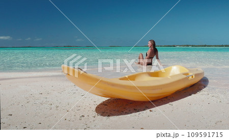 Woman relax sitting on beach, yellow kayak on sand, blue sky, turquoise water. Tanned young female on tropical island back view. Outdoor lifestyle travel on summer holiday vacation, active sport Woman relax sitting on beach, yellow kayak on sand, blue sky, turquoise water. Tanned young female on tropical island back view. Outdoor lifestyle travel on summer holiday vacation, active sport 109591715