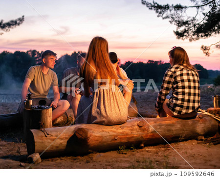 A company of young people sit around a fire at night on the banks of the river and relax in nature. Cooking food on a fire, outdoors 109592646
