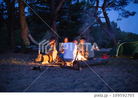 a company of friends have a rest in the evening on the banks of the river in nature. They fry sausages and talk. Cozy atmosphere, light from the fire. Summer rest, background 109592705