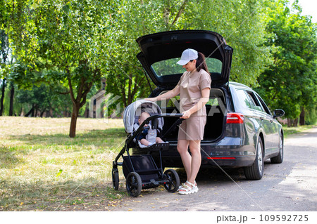 A beautiful girl puts a baby stroller into the open trunk of a car. Family car concept. Sunny weather in summer. 109592725